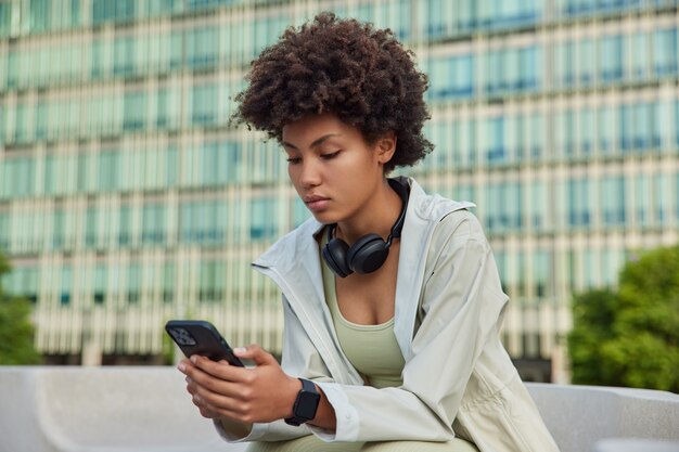 La mujer tiene el teléfono inteligente moderno comprueba la notificación comprueba la actividad durante el descanso después del entrenamiento navega por internet plantea contra el edificio moderno