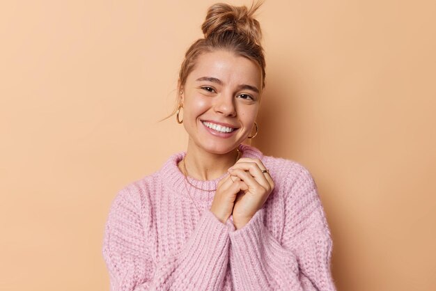 Una mujer tierna y de buen aspecto con el pelo peinado mantiene las manos juntas, sonríe suavemente y recibe cumplidos reconfortantes vestida con un suéter de punto informal aislado sobre un fondo beige. sentimientos agradables