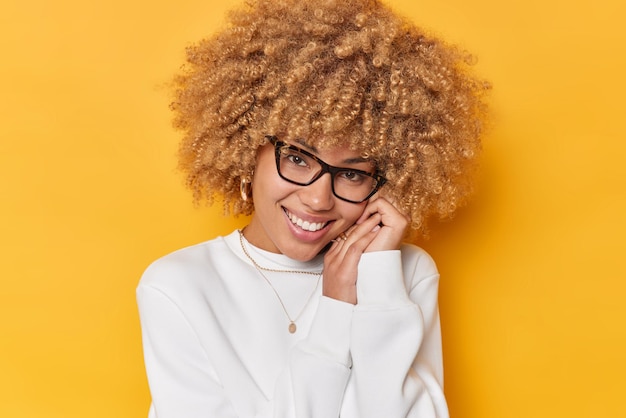 Mujer tierna encantadora mantiene las manos cerca de la cara mira suavemente a la cámara oye palabras agradables lleva gafas ópticas puente blanco casual aislado sobre fondo amarillo vivo. Concepto sincero de emotios