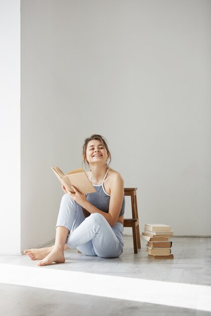 Mujer tierna hermosa joven que sonríe sosteniendo el libro que se sienta en piso sobre la pared blanca temprano en la mañana.