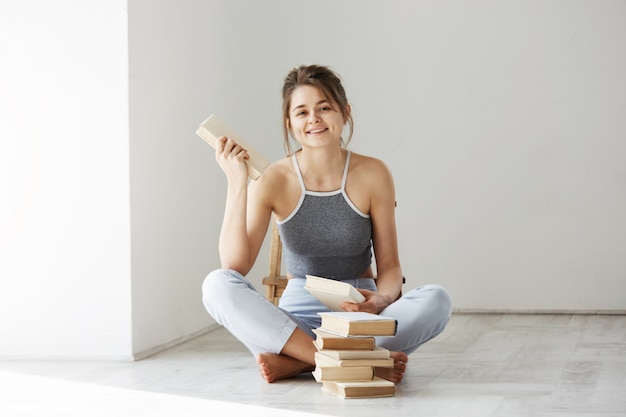 Mujer tierna hermosa joven que sonríe sosteniendo el libro que se sienta en piso sobre la pared blanca temprano en la mañana.