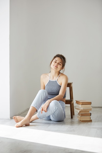 Mujer tierna hermosa que sonríe sentado en piso con los libros sobre la pared blanca.