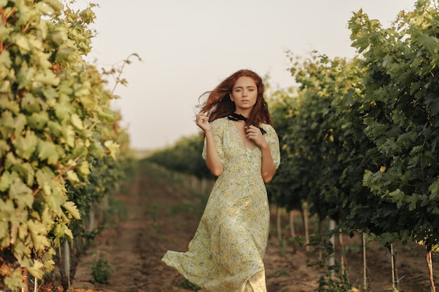 Mujer tierna con peinado ondulado rojo y vendaje negro en el cuello con un vestido largo y elegante de verano mirando al frente en un viñedo