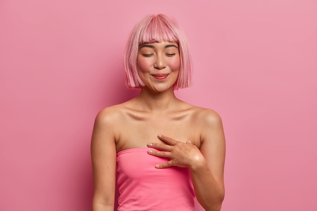 Foto gratuita la mujer tierna y tocada cierra los ojos, mantiene la mano cerca del corazón, feliz de recibir un regalo, recuerda una fecha hermosa, se ha teñido el cabello de rosa, se ha vestido de manera informal