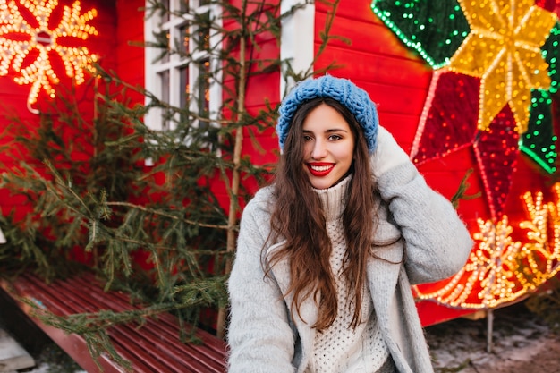 Mujer tímida con cabello largo castaño pasar tiempo en la feria de año nuevo y posando cerca de árboles verdes. Foto al aire libre de espectacular dama caucásica en abrigo gris de pie sobre adornos navideños rojos.