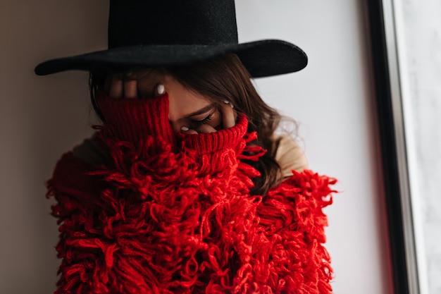 Mujer tímida se cubre la cara con las manos. Retrato de mujer con sombrero negro y suéter de punto rojo en sala blanca.