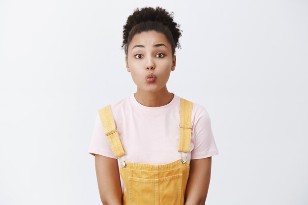 Mujer tímida esperando nerviosamente el primer beso, de pie con un lindo mono amarillo sobre una camiseta, doblando los labios para dar mwah, posando tierna y femenina sobre una pared gris