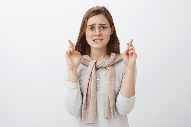 Mujer tímida, insegura e insegura preocupada con gafas y atuendo de moda mordiendo el labio inferior nerviosamente cruzando los dedos para la buena suerte esperando que suceda un milagro mientras reza para que el sueño se haga realidad