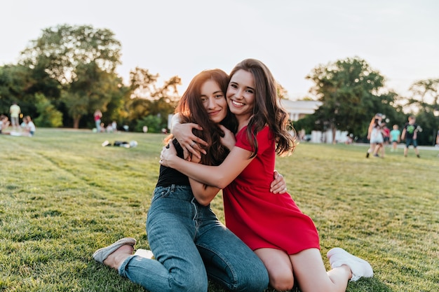 Mujer tímida en jeans abrazando a su amiga mientras está sentada en la hierba. Señoras de buen humor disfrutando del fin de semana de verano en el parque.