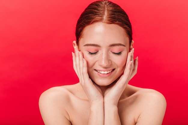 Mujer tímida sonriendo con los ojos cerrados Foto de estudio de una chica pelirroja tocando la cara con fondo rojo