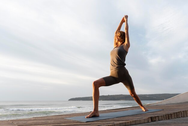 Mujer de tiro completo practicando yoga en la estera cerca del mar
