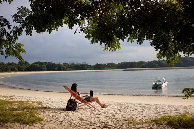 Mujer de tiro largo que trabaja en la computadora portátil en la playa