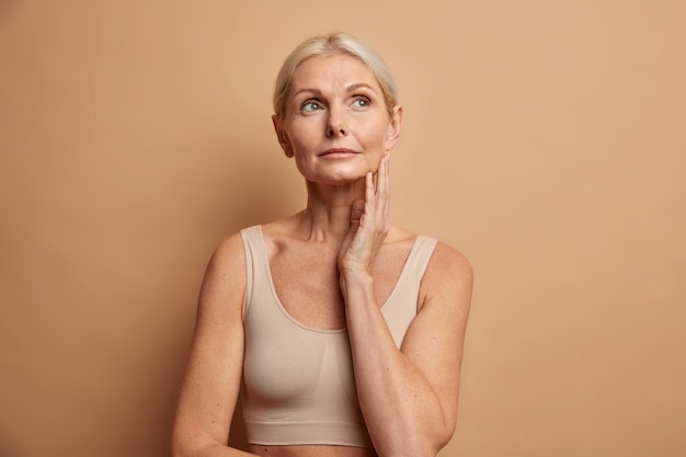 La mujer toca la piel después de aplicar la crema anti-edad concentrada arriba con expresión pensativa viste parte superior recortada aislado en marrón