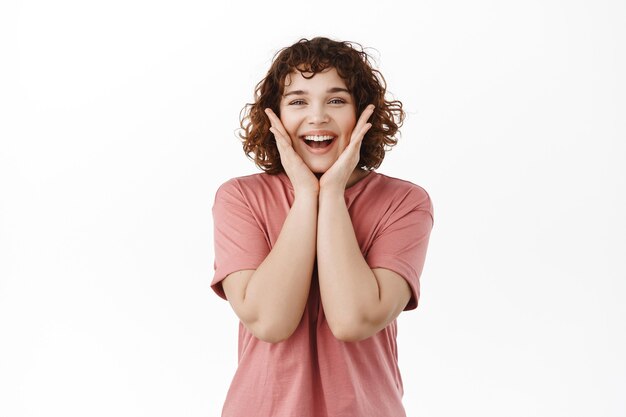 mujer, tocando mejillas y regocijándose, sorprendida y complacida por algo positivo y alegre, de pie sobre blanco.