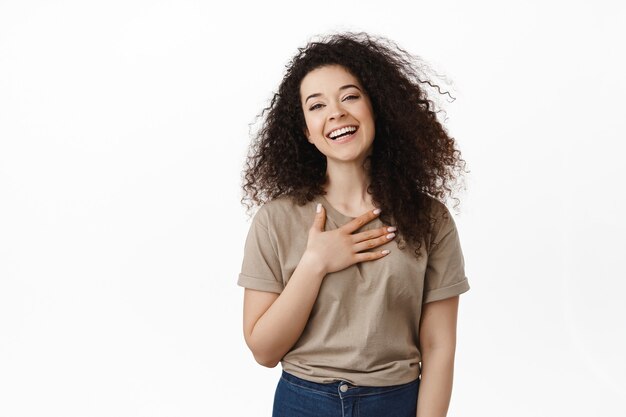 la mujer toma la mano en el corazón, riendo y luciendo natural, expresa gratitud, sintiéndose conmovida, de pie sobre blanco.