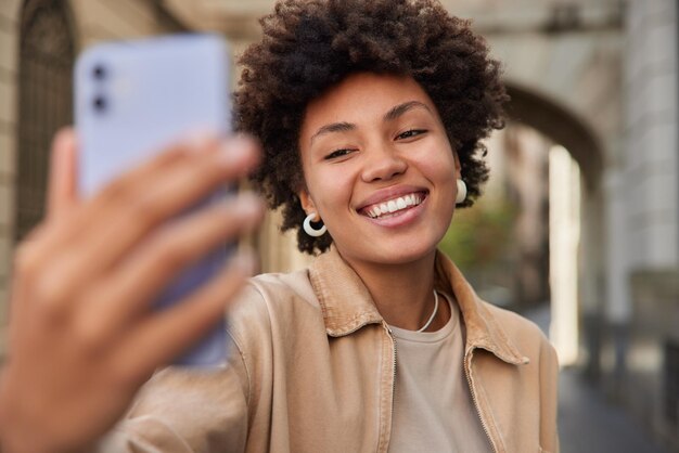 mujer se toma una selfie a través de un teléfono inteligente hace clic en las fotos de una cámara web celular hace que las fotos se publiquen en las redes sociales usa poses de ropa informal en la calle durante el día