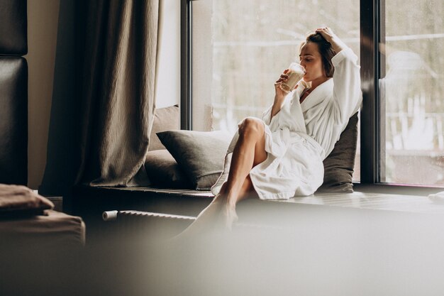 Mujer tomando café en bata de baño junto a la ventana en casa