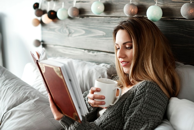 Mujer tomando café y leyendo un libro en la cama