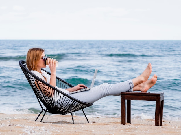 Mujer tomando café en la playa mientras trabajaba en la computadora portátil