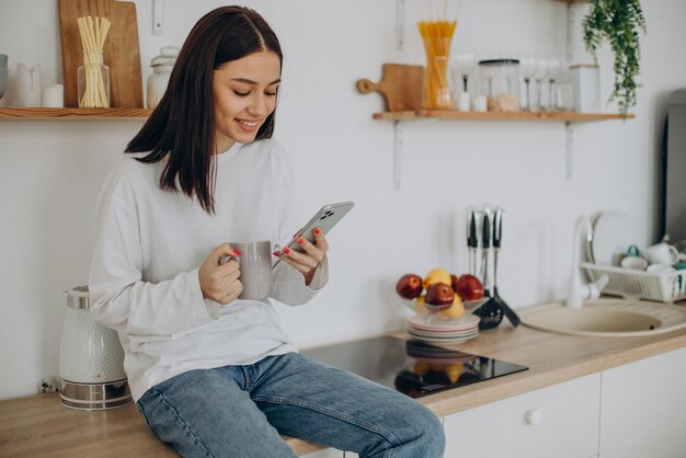 Mujer tomando café y usando el teléfono en la cocina