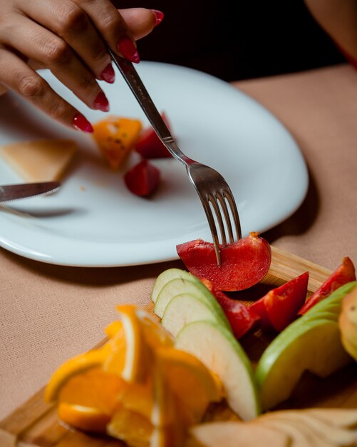 Mujer tomando ciruelas con carne de cerdo del plato de frutas con manzanas, naranjas, plátanos