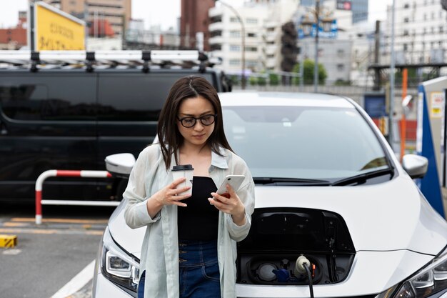 Mujer tomando un descanso para tomar café mientras su auto eléctrico se carga y usa un teléfono inteligente