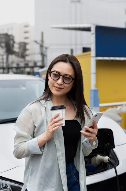 Mujer tomando un descanso para tomar café mientras su auto eléctrico se carga y usa un teléfono inteligente