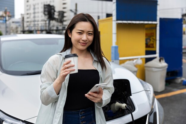 Mujer tomando un descanso para tomar café mientras su auto eléctrico se carga y usa un teléfono inteligente