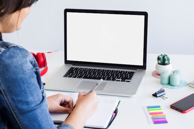 Mujer tomando notas cerca de computadora portátil y teléfono inteligente en la mesa