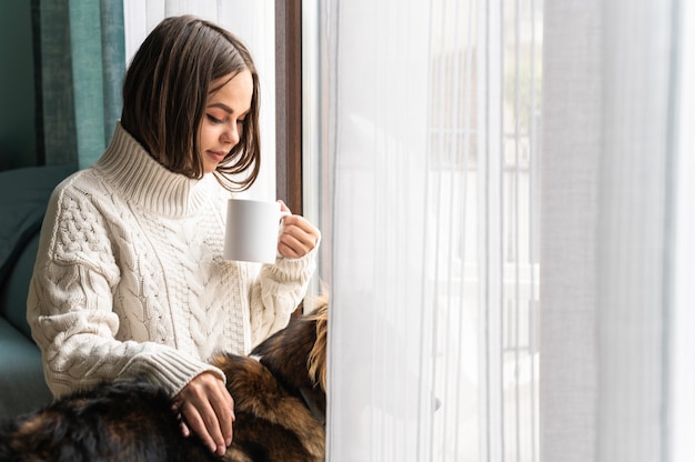 Mujer tomando una taza de café junto a la ventana en casa durante la pandemia