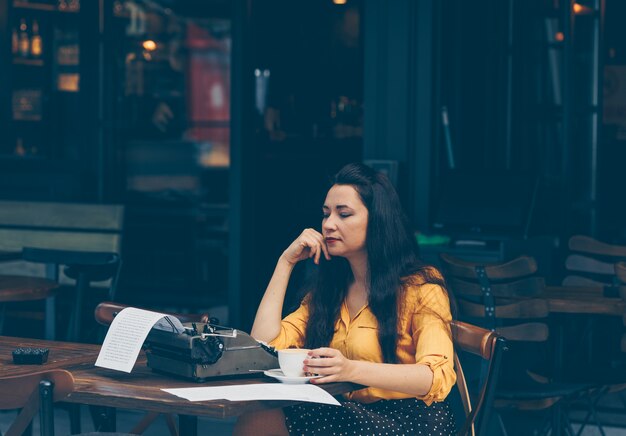 Mujer en top amarillo y falda larga sentada y tomando café en la terraza del café y mirando pensativo durante el día