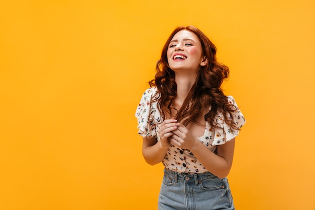 Mujer en top con estampado floral se ríe y se toca coquetamente su ondulado cabello rojo sobre fondo naranja.