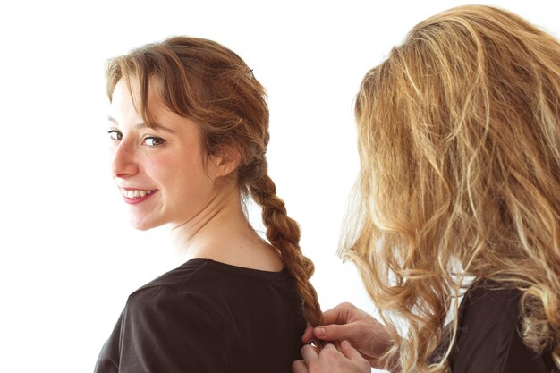 Mujer torciendo el pelo de la trenza de su hermana sonriente contra el telón de fondo blanco