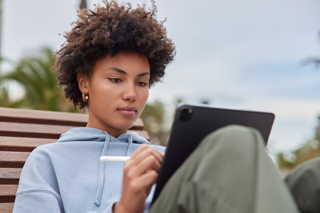 mujer trabaja en tableta hace diseño gráfico dibuja bocetos trabaja como ilustradora independiente vestida de manera informal plantea al aire libre utiliza bolígrafo electrónico para escribir