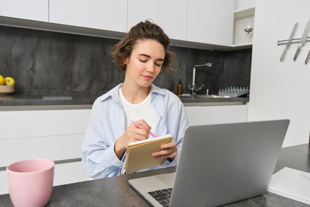 Mujer trabajadora hace notas escribe información en un cuaderno sentada con una computadora portátil en casa