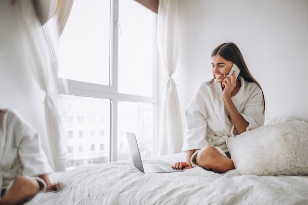 Mujer trabajando en la computadora en la cama y hablando por teléfono