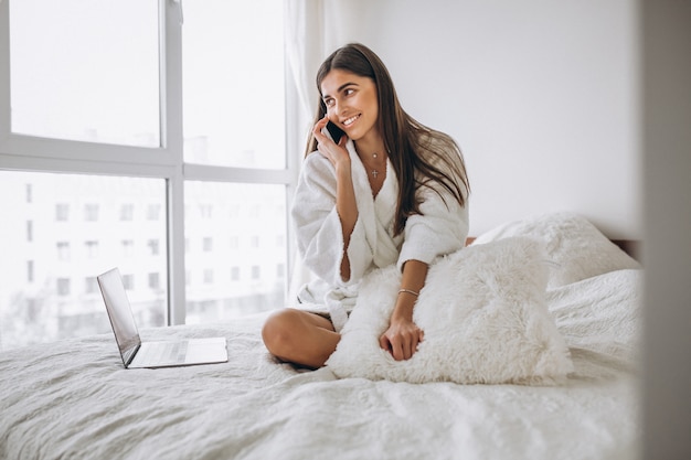 Mujer trabajando en la computadora en la cama y hablando por teléfono