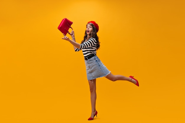 Mujer en traje elegante corriendo sobre fondo naranja. nd bolsa de sujeción. Chica joven sorprendida en boina y blusa a rayas posando.