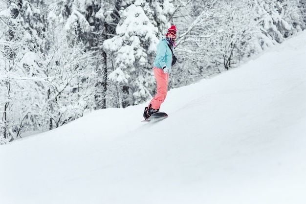 La mujer en traje de esquí mira sobre su hombro bajando la colina en su tabla de snowboard