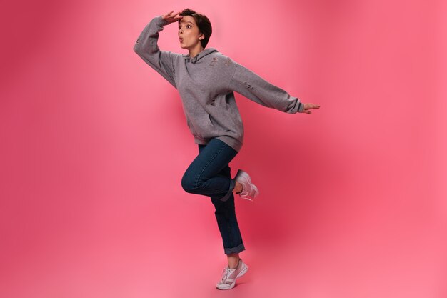 Foto gratuita mujer en traje de estilo callejero mira a distancia sobre fondo rosa. activo jovencita en jeans y sudadera con capucha saltando sobre aislados