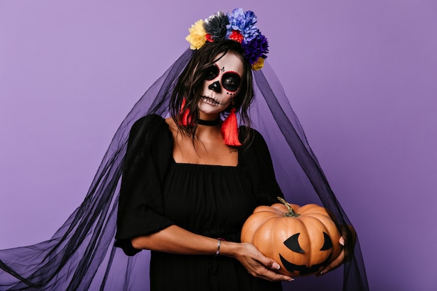 Mujer en traje de novia negro sostiene calabaza. Retrato de niña con flores en el pelo en la pared lila.