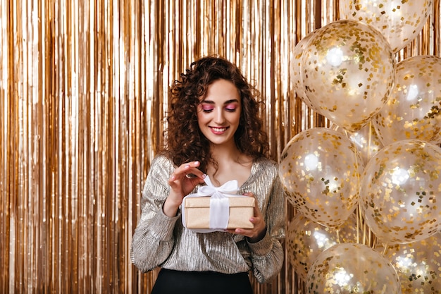 Mujer en traje plateado abre caja de regalo sobre fondo de globos