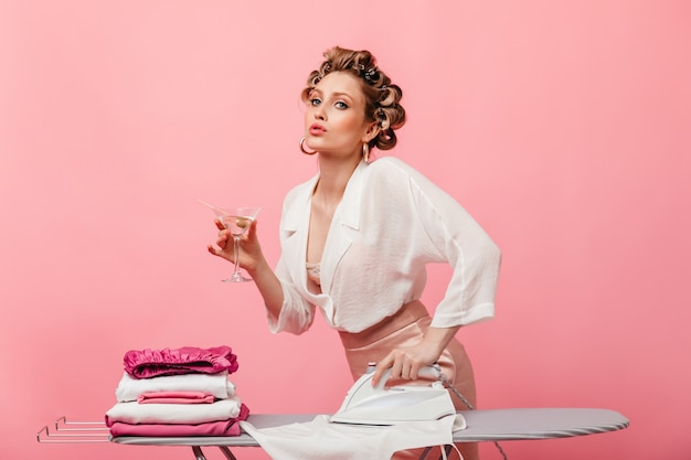 Mujer en traje de seda ligera posando en la pared rosa con copa de martini y planchando ropa