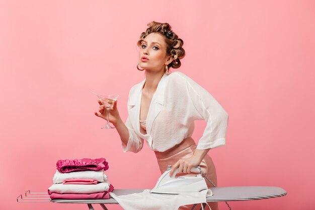 Mujer en traje de seda ligera posando en la pared rosa con copa de martini y planchando ropa