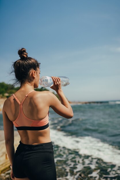 Mujer en traje de yoga bebiendo agua fresca de la botella después del ejercicio en la playa