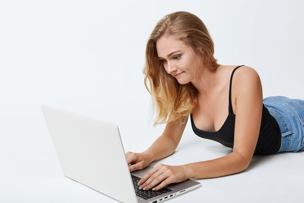 Mujer tranquila con cabello largo y rubio, acostada en el piso blanco frente a la computadora portátil abierta, enviando mensajes con amigos en las redes sociales, con una mirada concentrada y enfocada en la pantalla. Concepto de comunicación