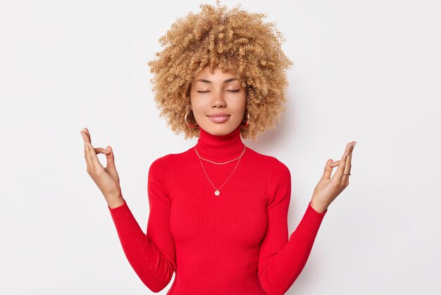 La mujer tranquila y concentrada con el pelo rizado mantiene los ojos cerrados y medita en el interior hace que el gesto de mudra viste un cuello de tortuga rojo informal aislado sobre fondo blanco que intenta aliviar el estrés. Pose de zen