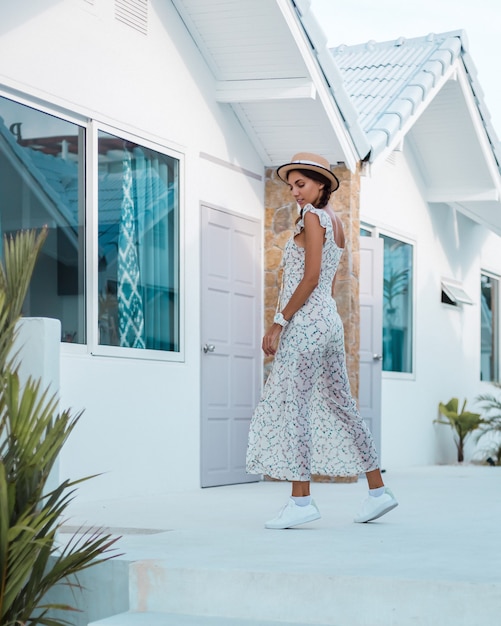 Mujer tranquila positiva en vestido ligero de verano, sombrero de paja, ubicación tropical