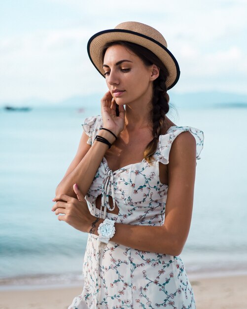 Mujer tranquila positiva en vestido ligero de verano, sombrero de paja, ubicación tropical