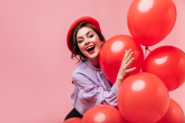 Mujer traviesa en boina roja se ríe y se divierte sobre fondo rosa con globos grandes.